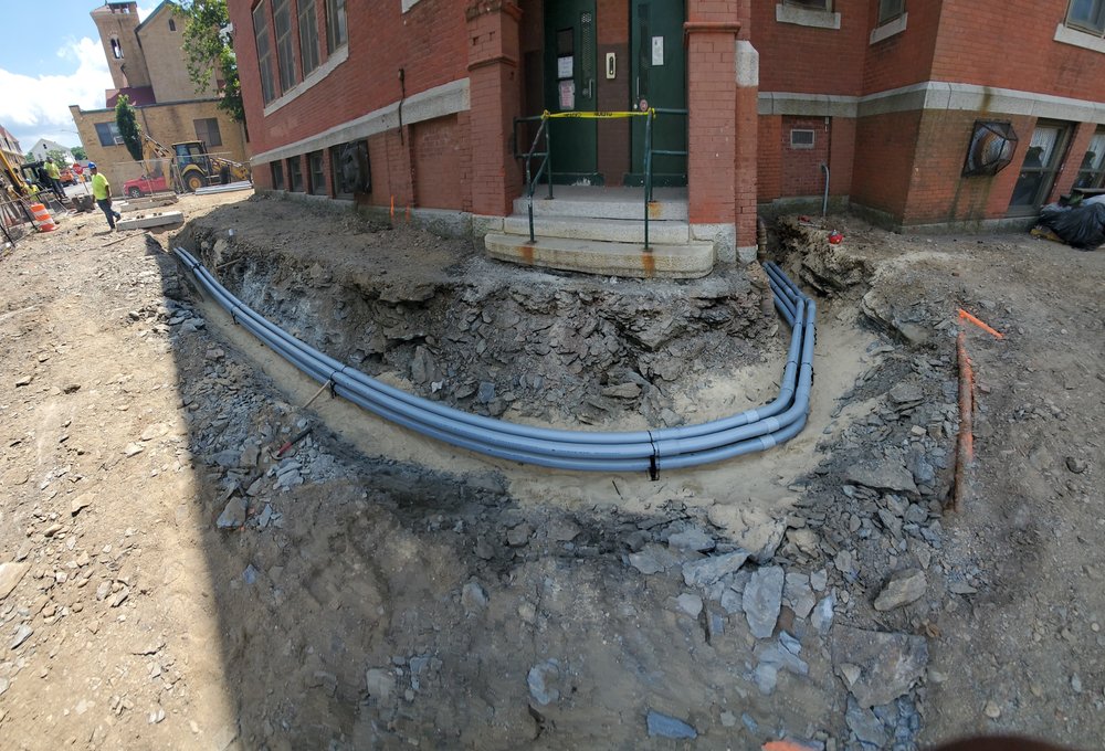 Gray conduit runs laid through an open construction trench beside a brick building.
